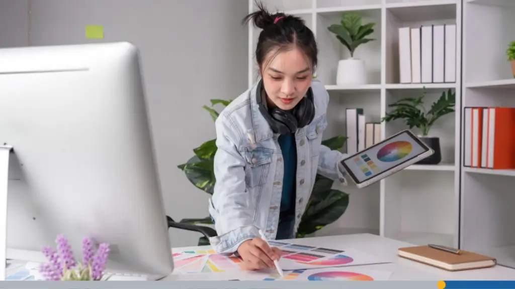 A young woman planning a logo using a tablet, desktop computer, and color swatch while designing a business card online.