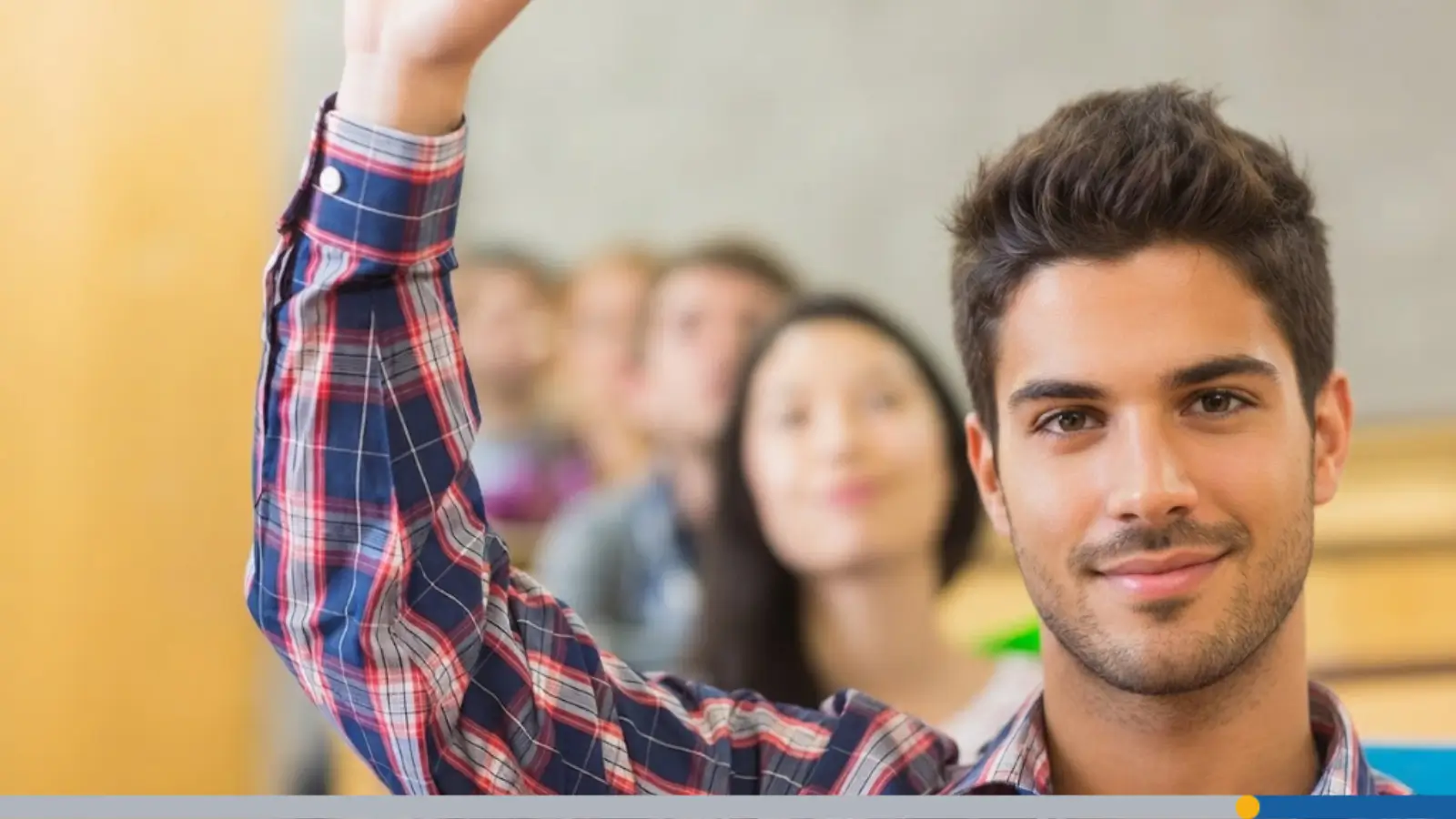 Young male student in a red and blue plaid shirt raising his hand while looking at the camera in a classroom, illustrating the ideal YouTube profile picture size.