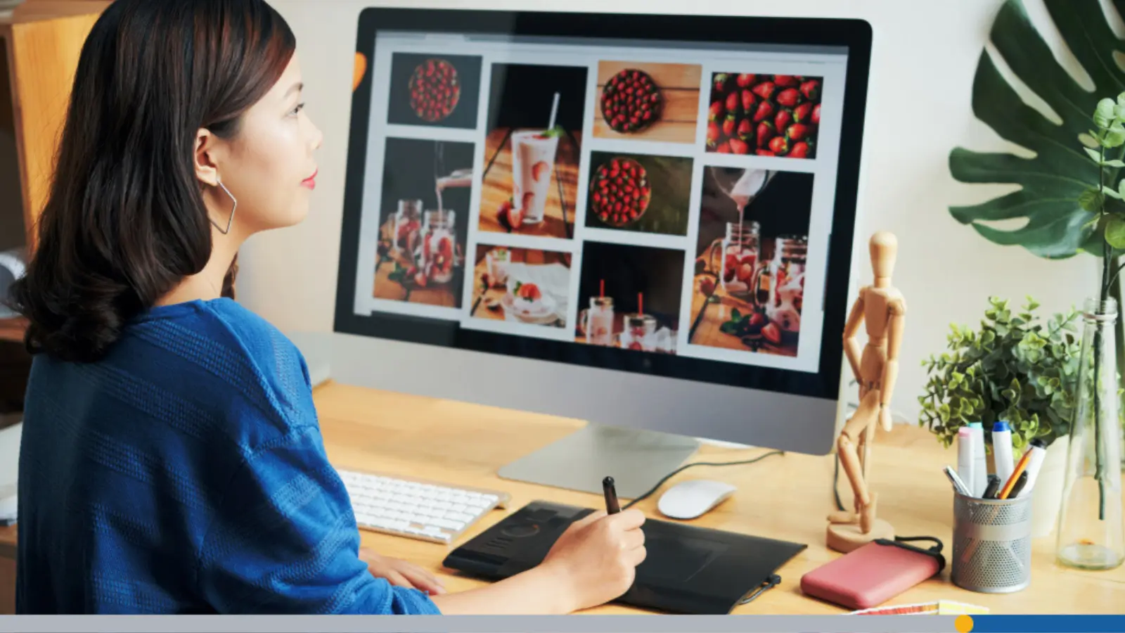 Side view of a designer at a desk using a stylus tablet and reviewing strawberry-themed food photos on a large screen, creating YouTube channel art at the recommended size.