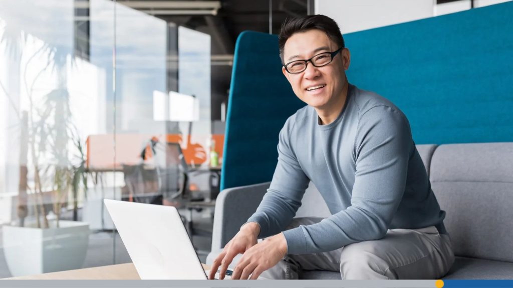 Professional working on his laptop computer, while sitting on a couch and smiling.