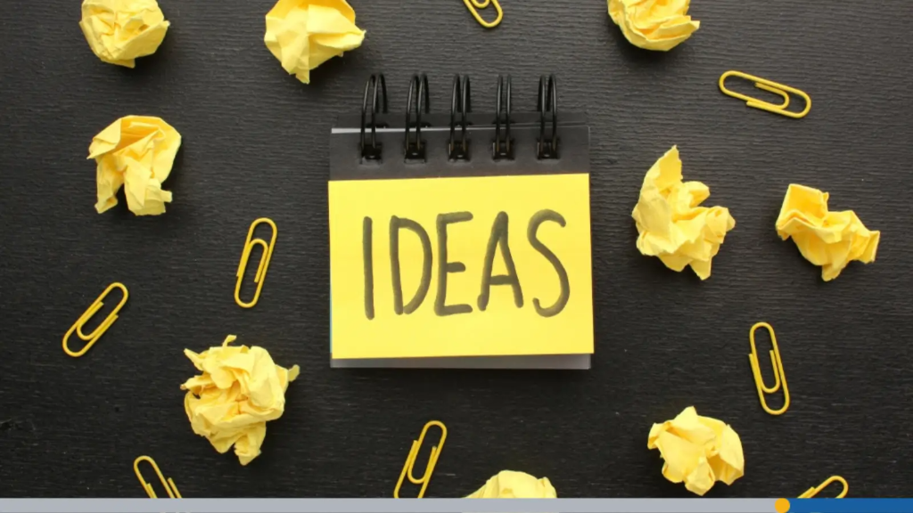 A small black notepad with a yellow sticky note reading "IDEAS" surrounded by crumpled yellow paper balls and paperclips on a dark surface, representing a search for the best business name ideas.