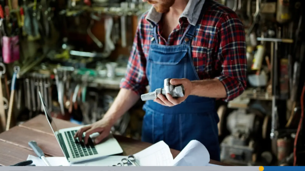 A tradesman in denim overalls and a plaid shirt holding a metal pipe fitting while typing on a laptop at his workbench, looking up how to pick a business name.