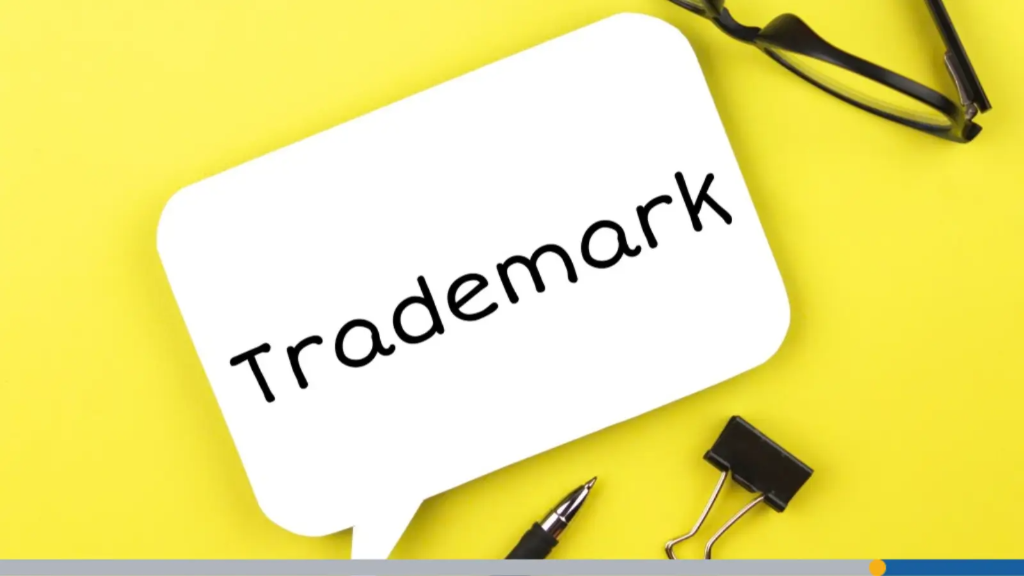 A white speech bubble note reading "Trademark" placed on a bright yellow background alongside eyeglasses, a pen, and a binder clip, showing a key step in how to choose a business name.