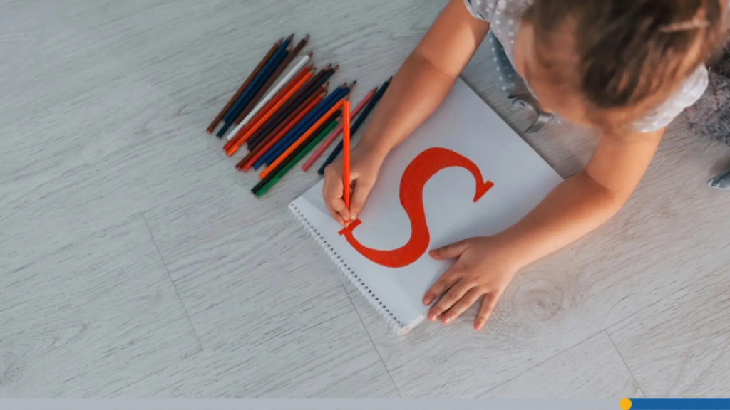 A young child lying on the floor coloring a large red letter "S" inside a spiral sketchbook next to colored pencils, illustrating creative ways on how to pick a business name.