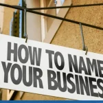 A white hanging shop sign mounted on a textured building wall featuring bold black text that reads "HOW TO NAME YOUR BUSINESS".