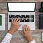 Man at a desk with a laptop, notebook, camera, phone and coffee