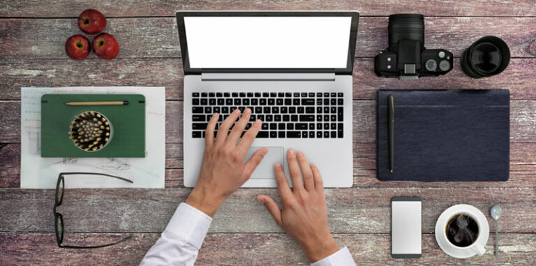 Man at a desk with a laptop, notebook, camera, phone and coffee