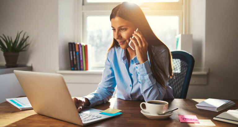 woman sitting at her desk on her phone looking at laptop